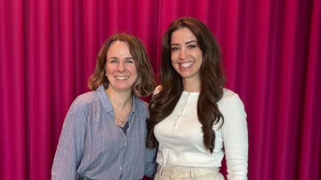 Cariad Lloyd on the left with shoulder length light brown hair and a light blue stripey shirt and Dr Sasha Hall with long dark wavy hair wearing a white button up long sleeve top. They are standing close side by side in front of a red curtain
