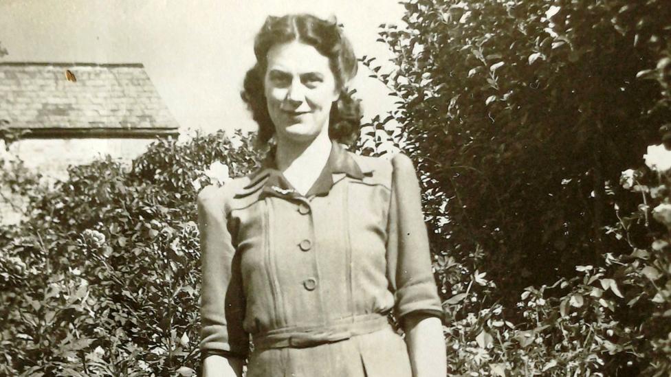 A black and white photo of a woman smiling at the camera in front of bushes