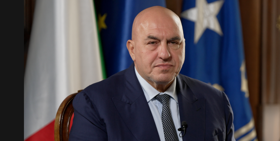 Guido Crosetto poses wearing a suit in front of the Italian and EU flags. 