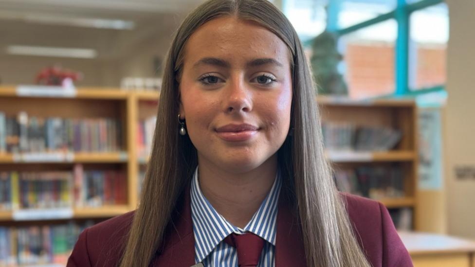 A teenage girl with long light brown hair looks at the camera. She has a burgundy blazer blue and white stripery shirt and burgundy tie
