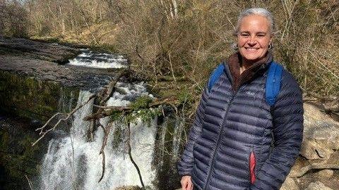 Eleanor sharsharasi yonida turibdi. Uning oq sochi orqaga bog'langan, harbiy ko'k palto kiygan. Kamerga tabassum bilan qarayapti.