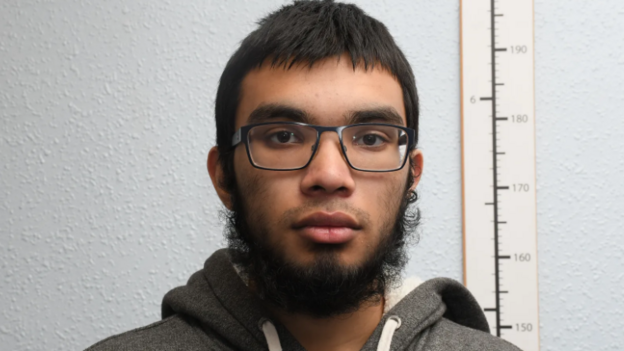 Yusuf Shah, a young man with a beard, short dark hair and black glasses, stands against a plain wall next to a long ruler. He is wearing a grey hoodie.