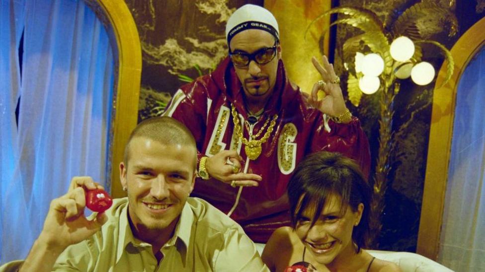 Мужчина в спортивной куртке в стиле Али Джи показывает два знака «ОК» руками, а мужчина и женщина улыбаются в камеру и позируют с красными носами Comic Relief в руках.