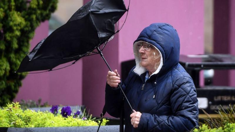 A woman struggling with her umbrella turned inside out - she is wearing a blue puffy jacket with the hood up