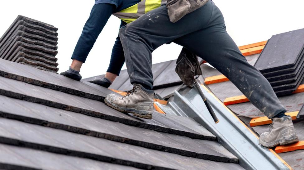 Lower half of a workman standing on a roof with a chimney visible. Some tiles are not yet laid on the roof structure.