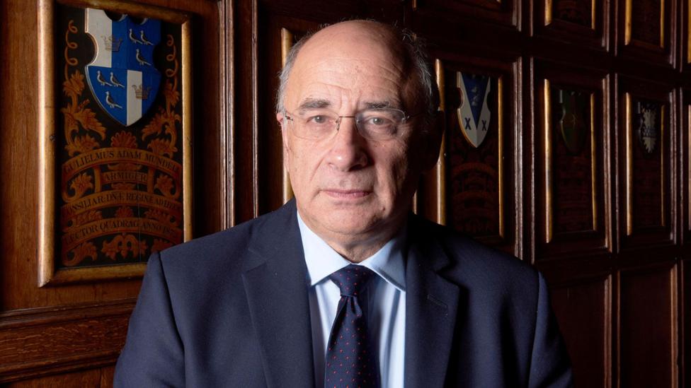 A head-and-shoulders portrait of Sir Brian Leveson, a white man with rimless glasses and grey hair at the sides of his head, wearing a navy suit with a light blue shirt and navy tie. He is standing in a wood-panelled room with heraldic designs behind him and is lit by soft light coming from the left of the picture.