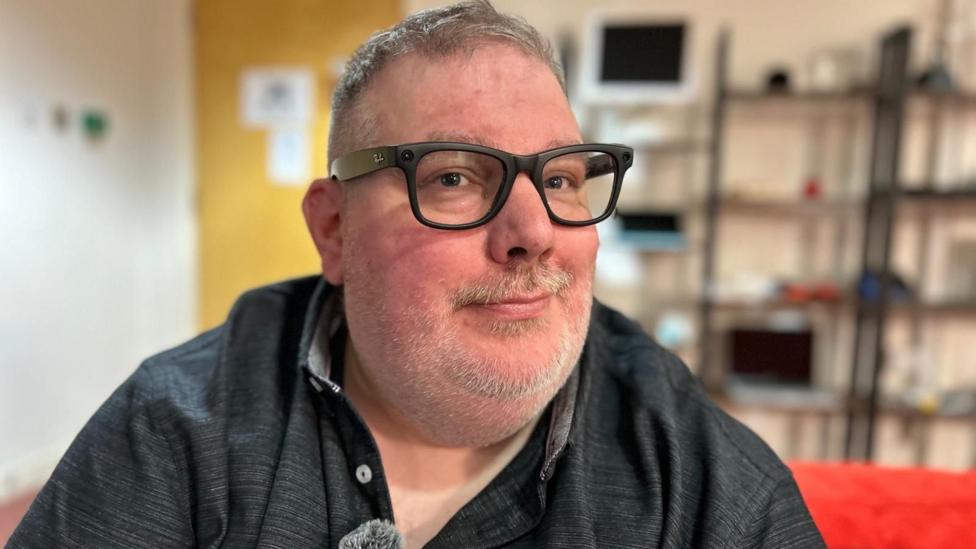 Close-up portrait of Gordon Brown wearing black-framed glasses and a dark collared shirt, seated indoors with shelving units, computer equipment and a red beanbag blurred in the background.