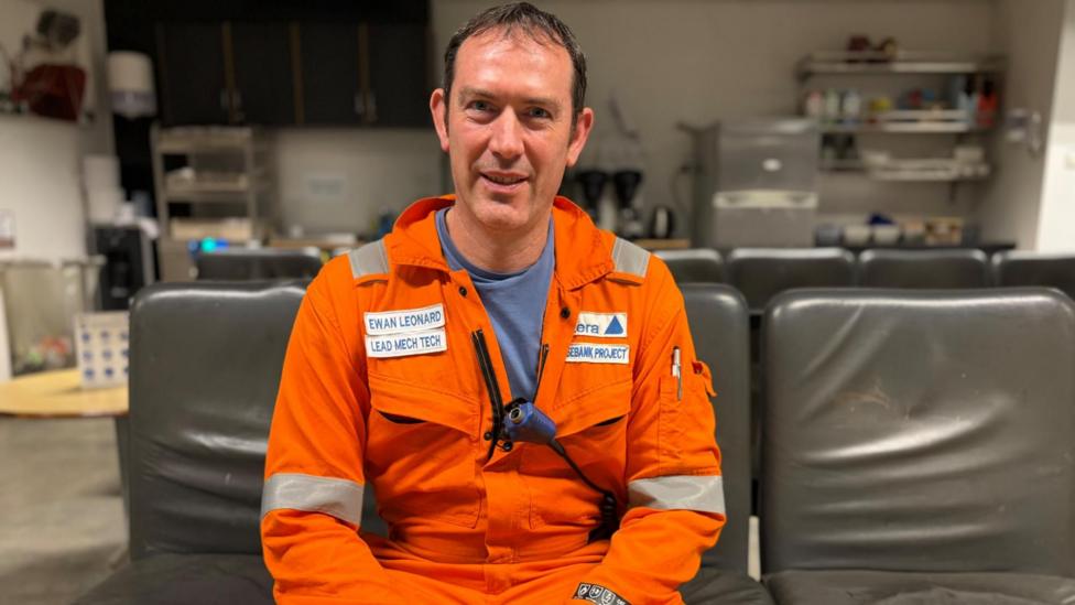 Ewan Leonard is sitting down on a grey seat. He has dark, receding hair and is wearing an orange boiler suit with his name on it. He is smiling.