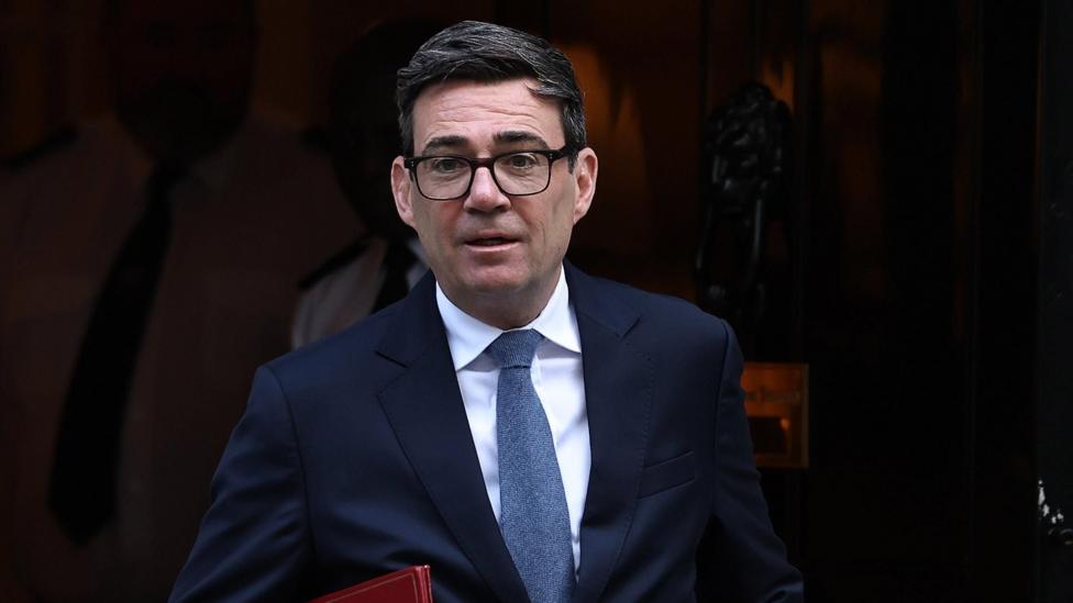 Andy Burnham leaving Downing Street. He is wearing a dark blue suit, blue tie and glasses. He is carrying a red folder.