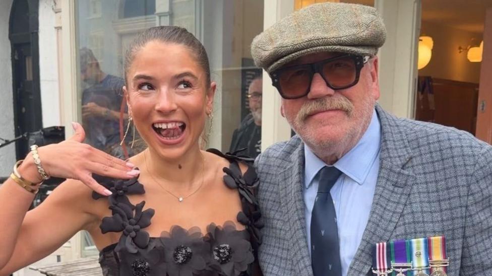 Ray Winstone has a big moustache, thick rimmed glasses and a blue blazer with military medals on the lapel, he is seen with Molly Ridge who is wearing a black dress made of fabric flowers and acts shocked to see the film star