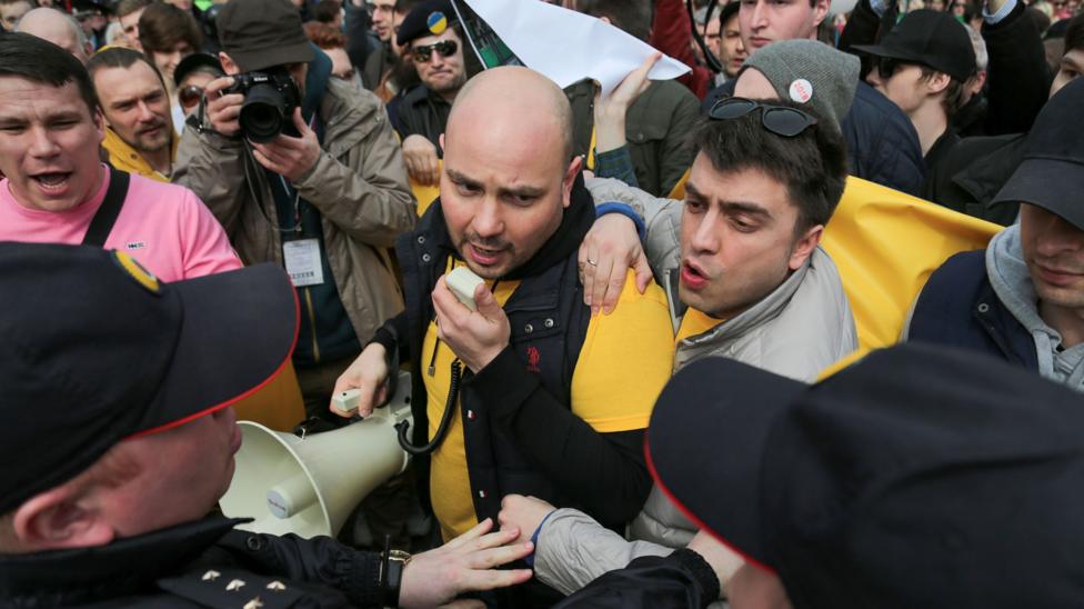 Andrei Pivovarov is at the centre of the shot speaking into a megaphone and is surrounded by other protestors. Russian police are approaching him and reaching towards him. 