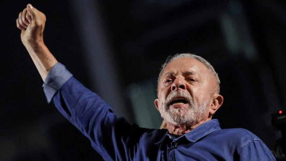 President Lula stretches his fisted right arm as he addresses supporters. He's wearing blue shorts and has a grey beard and short hair.