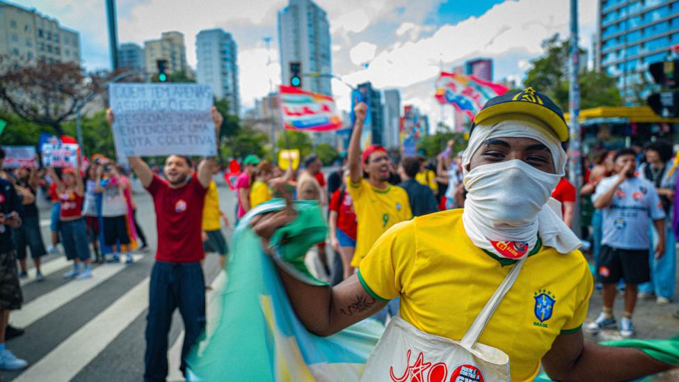 Yuzini oq mato bilan yopgan, sariq Braziliya futbolka kiygan yosh yigit Braziliya bayrog‘ini silkitmoqda. Orqa fonda namoyishchilar bayroqlar va plakatlar ushlab turishgan.