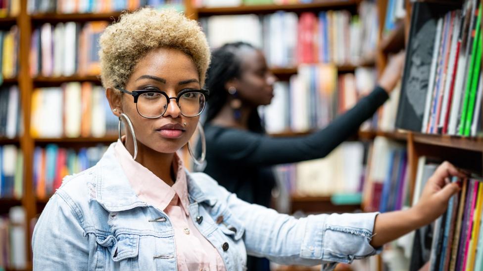 A  female university student in a library wearing a demin jacket looking towards the camera with her hand on a book