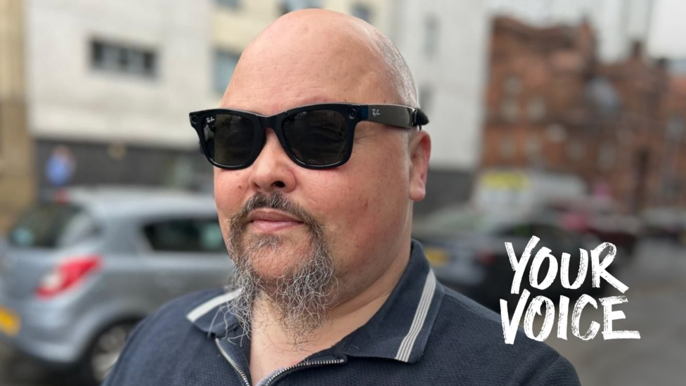 Portrait of Fraser Fleming wearing dark sunglasses and a navy zip-neck polo shirt, standing on a wet city street with parked cars and buildings blurred in the background.