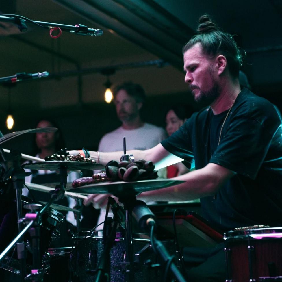 Мужчина с пучком волос сидит за ударной установкой. На нём чёрная футболка, он на сцене с фиолетово-синим освещением.