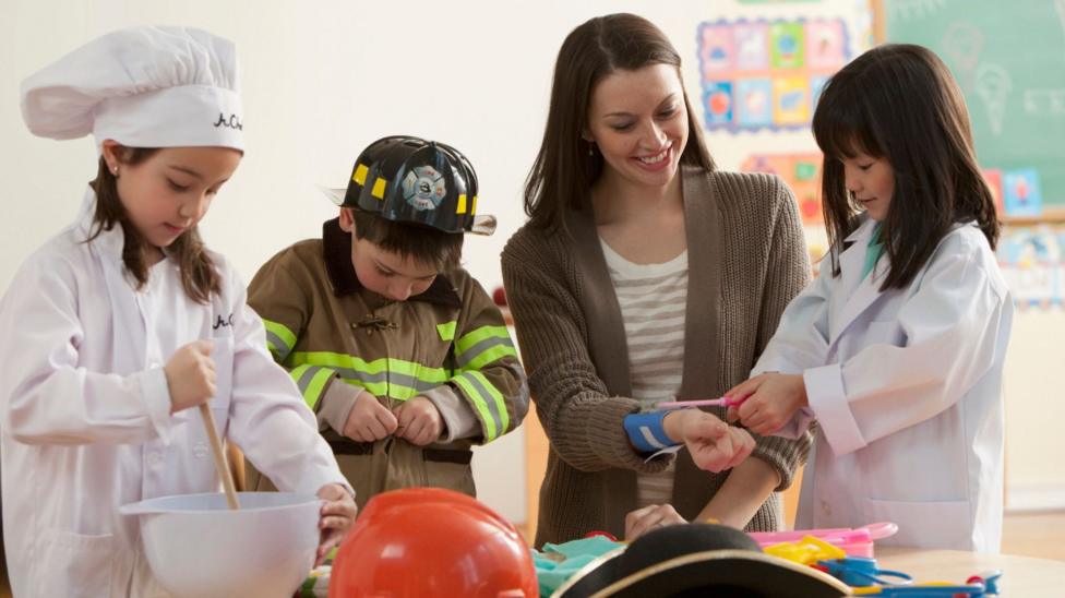 Three young children of primary school age are dressed up as a chef, a fireman and a nurse alongside their teacher in a classroom setting.