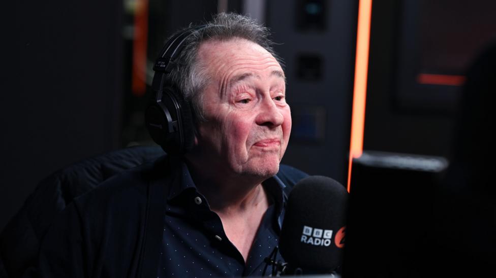 A man with grey hair and wearing a large set of headphones sits behind a BBC Radio 2 microphone.