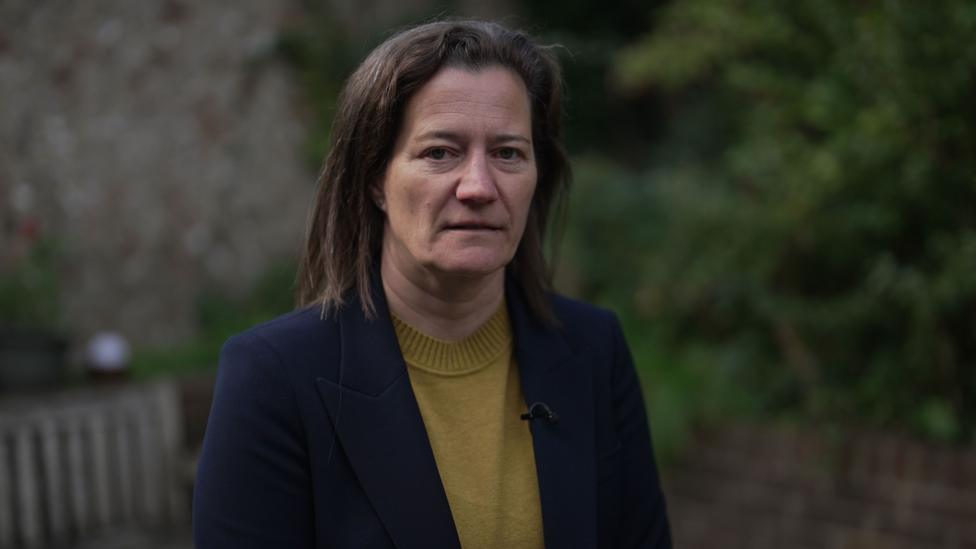 Dr Sarah Redvers outside in a garden wearing a navy blazer and mustard jumper. She has shoulder length brown hair