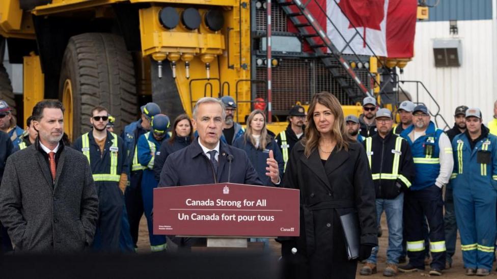 Mark Carney speaks at a podium with a sign that reads 'Canada Strong for All'. Next to him, in a black suit, is Danielle Smith. Behind them a crowd of workers stand in front of heavy equipment and a Canadian flag.