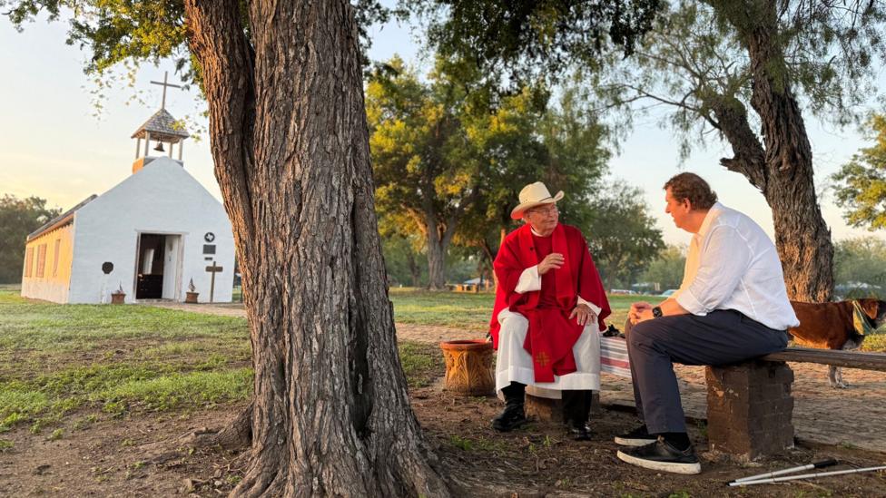 Image shows Father Roy Snipes sitting outside the small, white chapel with the BBC's Gary O’Donoghue