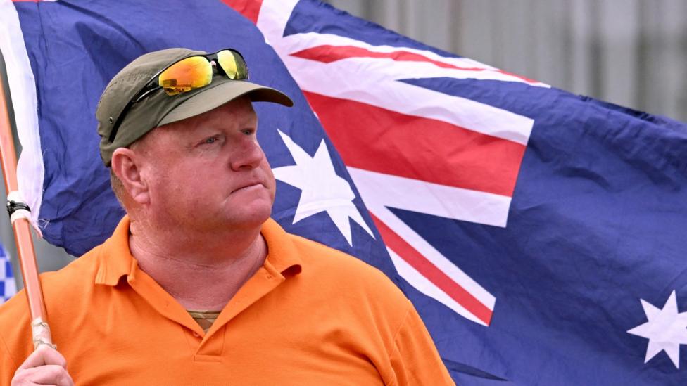 A man wearing an orange t-shirt reading 'Wangaratta this is not us!!' waves an Australian flag