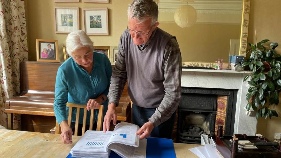 Bern Rogers and her husband John look through a folder of documents on the kitchen table in their house with a piano, marble mantelpiece and large mirror in the background.