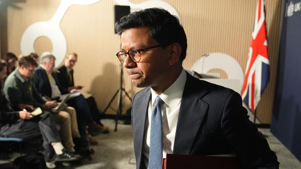 Kanishka Narayan podiumdan nutq so'zlagach ketayotgan holda: ko'k galstukli kostyumda, orqada Buyuk Britaniya bayrog'i va ko'k fon, orqasida matbuot vakillari