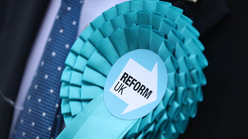 A light blue Reform UK rosette pinned on a person wearing a suit and a dark blue tie.