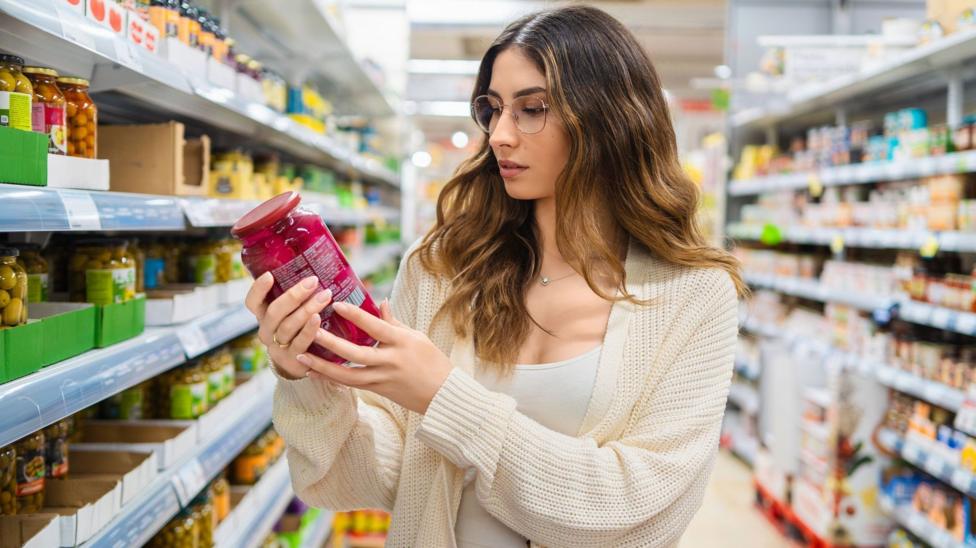 Молодая женщина в очках рассматривает банку в проходе продуктового магазина. Она сосредоточенно читает этикетку, вокруг — разнообразные продукты