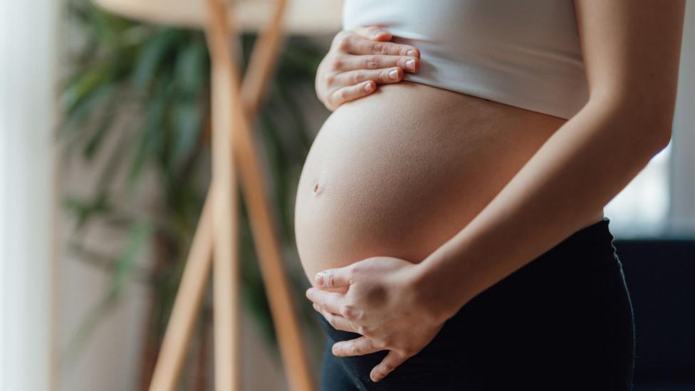 A pregnant woman places her hand on the top and bottom of her baby bump. She is wearing a white cropped top and her pregnant belly is centre of the frame. She is wearing black leggings and is in a living room.