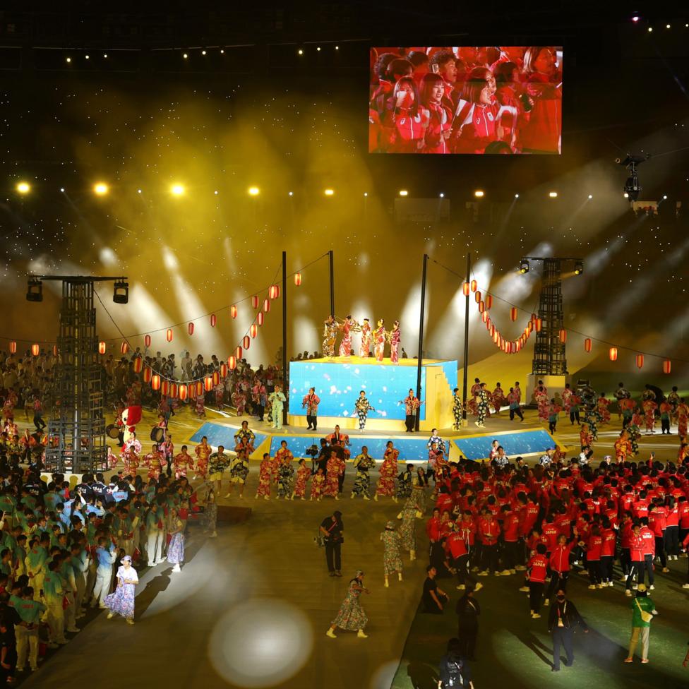 Performers in colourful traditional costumes dance around a central stage decorated with hanging red lanterns during the Aichi-Nagoya Asian Games closing ceremony, while groups of athletes gather on the arena floor under bright gold and white spotlights. A giant video screen above the stage shows participants in red uniforms, and small lights twinkle in the darkened stands.