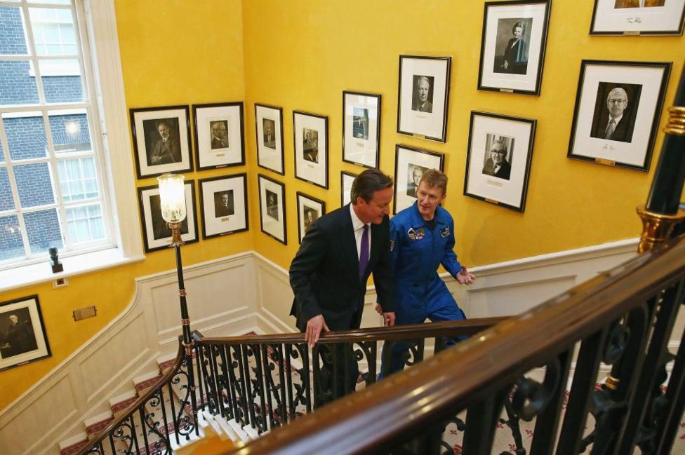 British astronaut Major Tim Peake walks up the stairs with then-Prime Minister David Cameron in 10 Downing Street in London. The staircase winds around a central lobby, with portraits on a yellow wall.