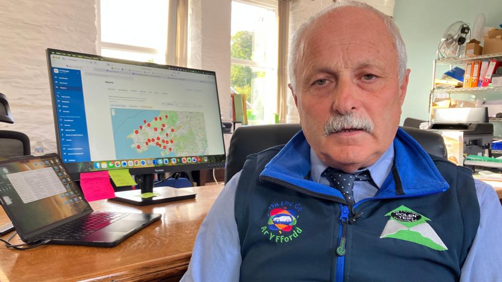 A photo of a man with a moustache wearing a dark blue bodywarmer. He's sitting in an office with his back to a desk which has a computer monitor and laptop on it.