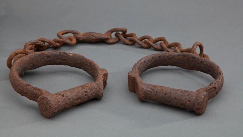 Two iron shackles consisting of iron loops joined together by a length of iron chain. The iron is heavily rusted. These would have been used to restrain enslaved African people forcibly transported to North America and the Caribbean.