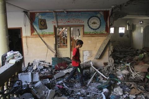 A boy in a red t-shirt walks through piles of rubble and strewn wires inside a school building. He is walking in front of a wall with a blue mural.