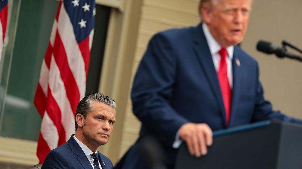 Pete Hegseth o‘tirgan holda, prezident Tramp esa ko‘k kostyum va qizil galstukda tribunada turibdi