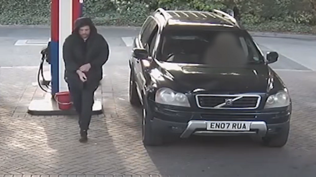 Qora pufakça kurtka kiygan, kapyuşonini kötargan va qora şim kiygan oq tanli erkak benzin quyiş maydonidan qora rangli Volvo XC90 universal avtomobilidan uzoqlaşib yurib ketmoqda.