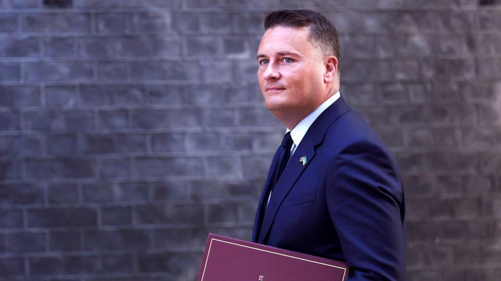 Wes Streeting walks in front of the Downing Street building facade carrying a binder that says 'Secretary of State for Defence' on the front, in September.