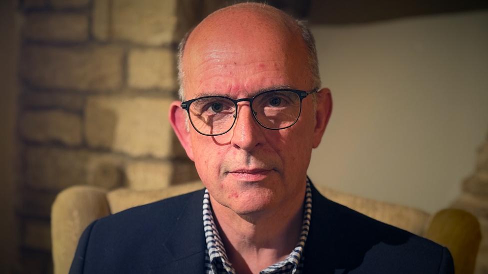 A head and shoulders shot of Andrew Coles sat on a chair in his living room. He is wearing spectacles, a shirt and a blue jacket. He has a bald head and greying hair.