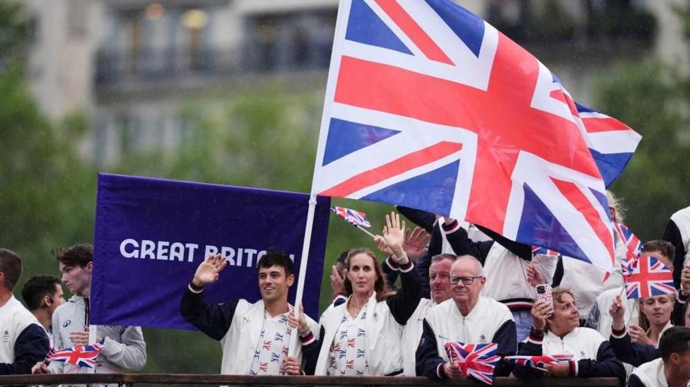 Tom Deyli va Team GBning boshqa a’zolari Parijdagi qayiqdan ulkan Union Jack bayrog‘ini ko‘tarib qo‘l silkitmoqda, ular orqasida binafsha mato ustidagi Buyuk Britaniya banneri ko‘rinib turibdi