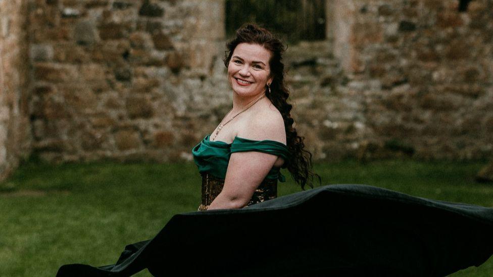A woman with long brown hair in an off-the-shoulder green dress and dark flowing skirt stands on a grassy lawn in front of a stone building. The skirt billows outward in motion, a stone wall forms the backdrop.