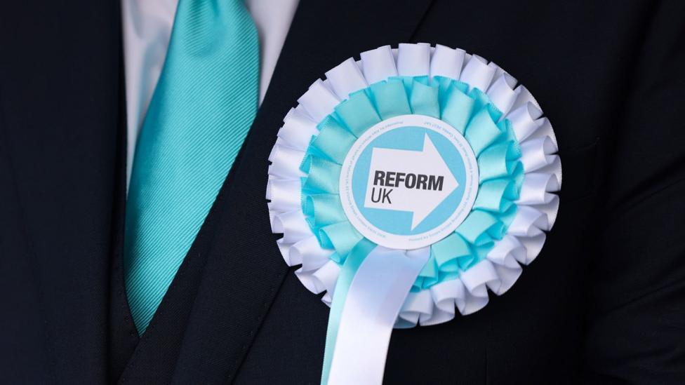 An anonymous person in a suit and a blue tie wears a Reform UK rosette