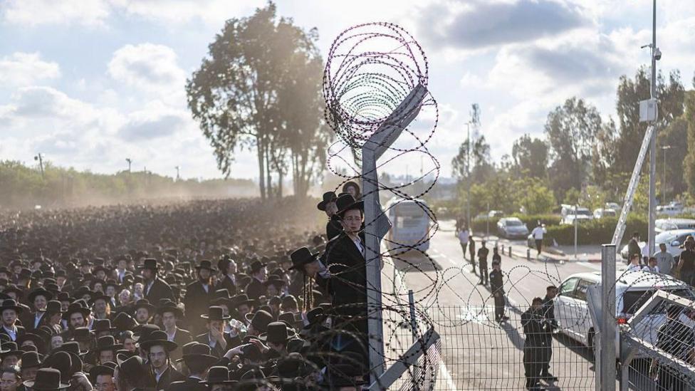 Hundreds or thousands of ultra-Orthodox jews stand beside barbed wire fencing.