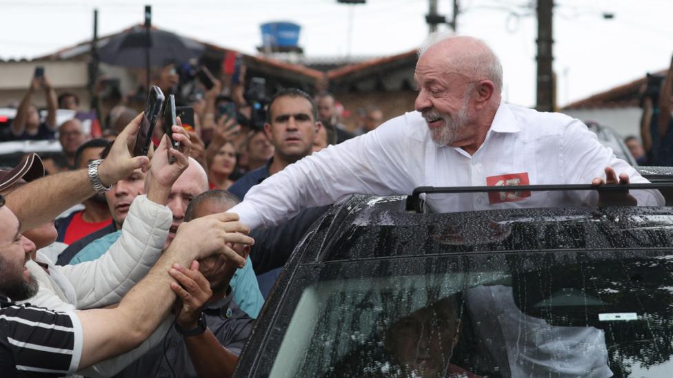 Oq soqolli, oq tugmali ko‘ylakdagi Lula yomg‘ir tomchilari tushgan avtomobil lyukasidan chiqib turib tabassum qilmoqda va qo‘llarini cho‘zib, shu lahzhani smartfonlarida videoga olmoqchi bo‘lgan tarafdorlari bilan qo‘l siqishga intilmoqda. Voqea gavjum ko‘chada bo‘lib o‘tmoqda. Uning orqasida xavotirli ko‘ringan bir erkak (xavfsizlik xodimi?) diqqat bilan kuzatmoqda.