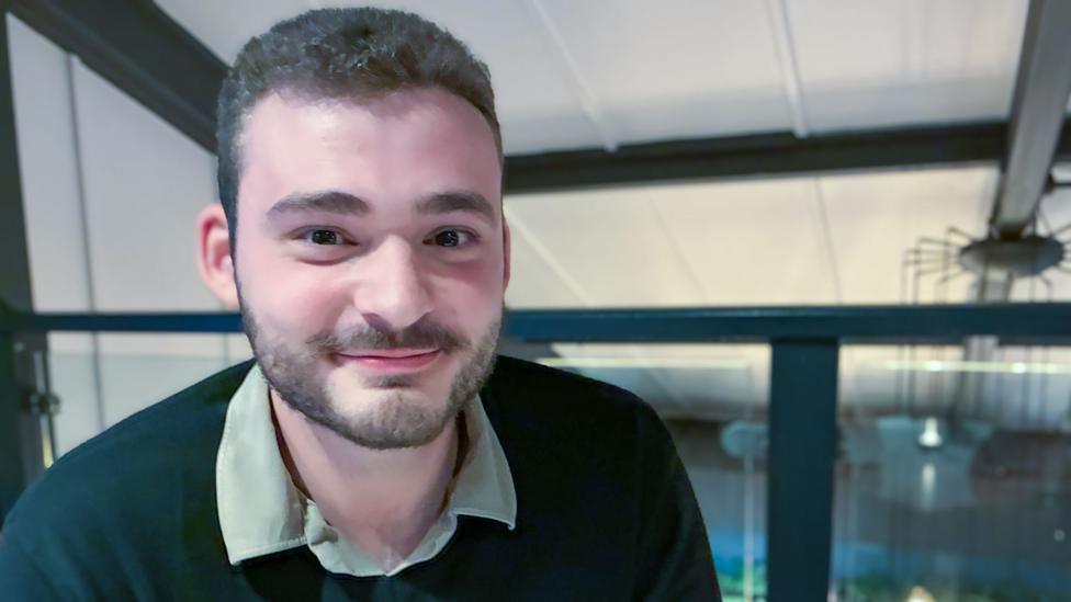 Edward, wearing a shirt and jumper, sits in a bar
