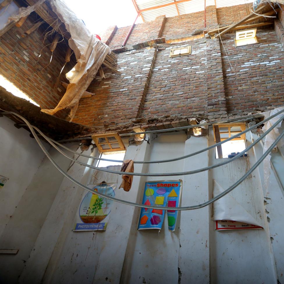Interior of a damaged classroom with part of the roof collapsed, exposing brick walls and open sky above. Twisted metal ceiling supports hang across the room, while educational posters, including one on photosynthesis and another showing basic shapes, remain attached to stained white walls.