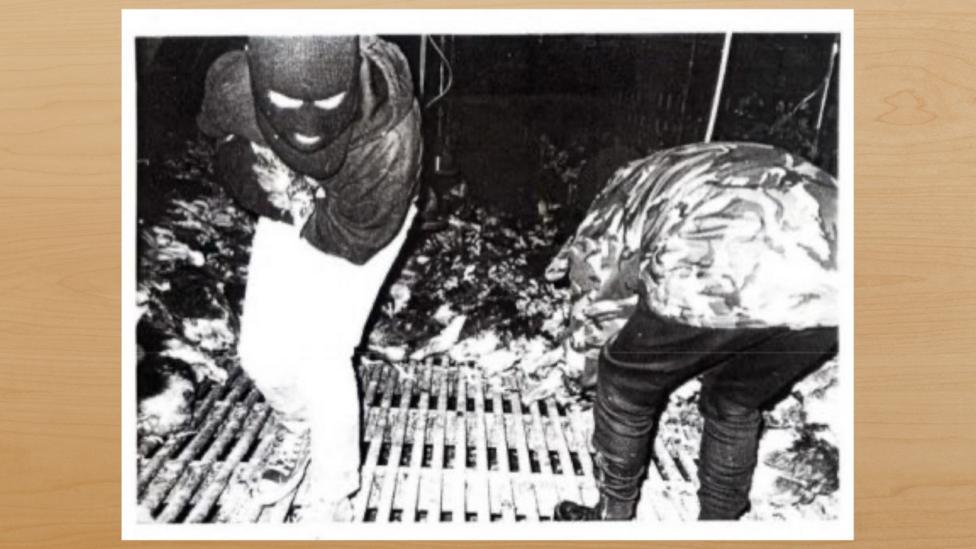 A black and white image from the inside of a laying hen unit. Two people are standing on the slatted floor in the middle. One is leaning over. The other, Jessica, is wearing a balaclava and clutching a chicken to her chest.