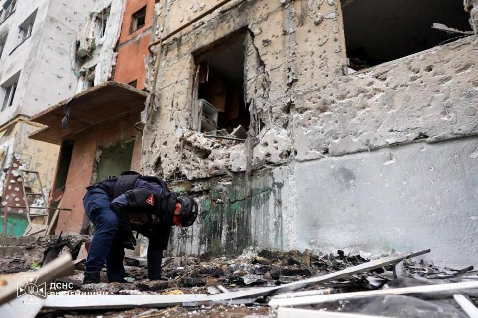 An official, wearing a helmet and protective gear, sifts through rubble. There is a heavily damaged building behind him.