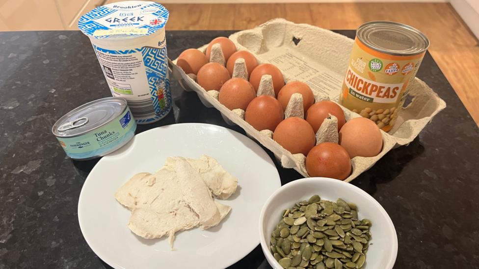 The photo shows a tin of tuna, a plate of cooked chicken, a bowl of pumpkin seeds, a tub of greek yoghurt, a carton of eggs and a can of chickpeas laid out on a black kitchen work surface. You can see a wooden floor in the background.
